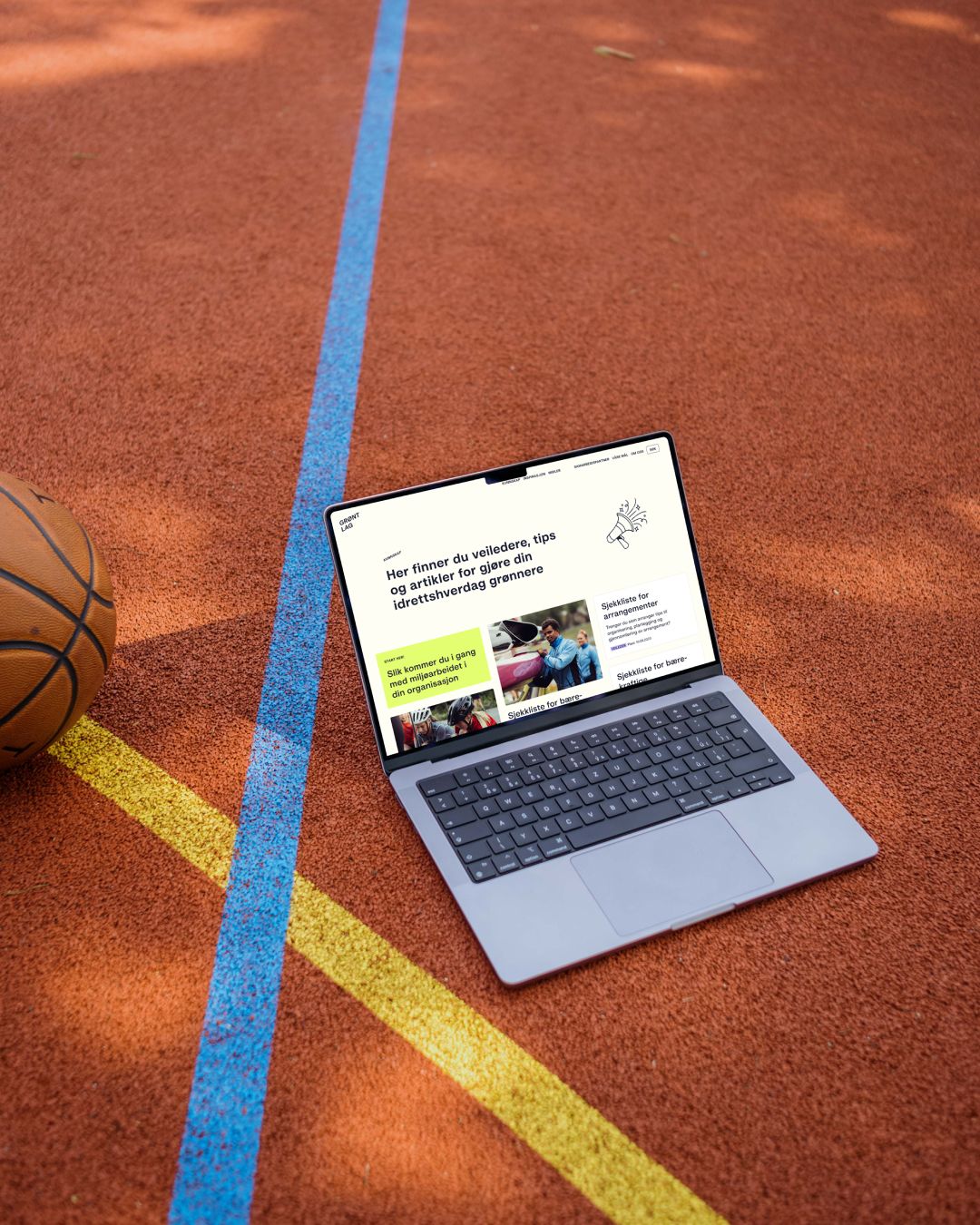 En bærbar PC med grontlag.no åpen står på en basketballbane ved siden av en ball.